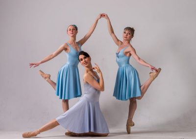 Trio of ballerinas in light blue and lavender; two dancers en pointe framing the third dancer who is kneeling with one leg out along floor