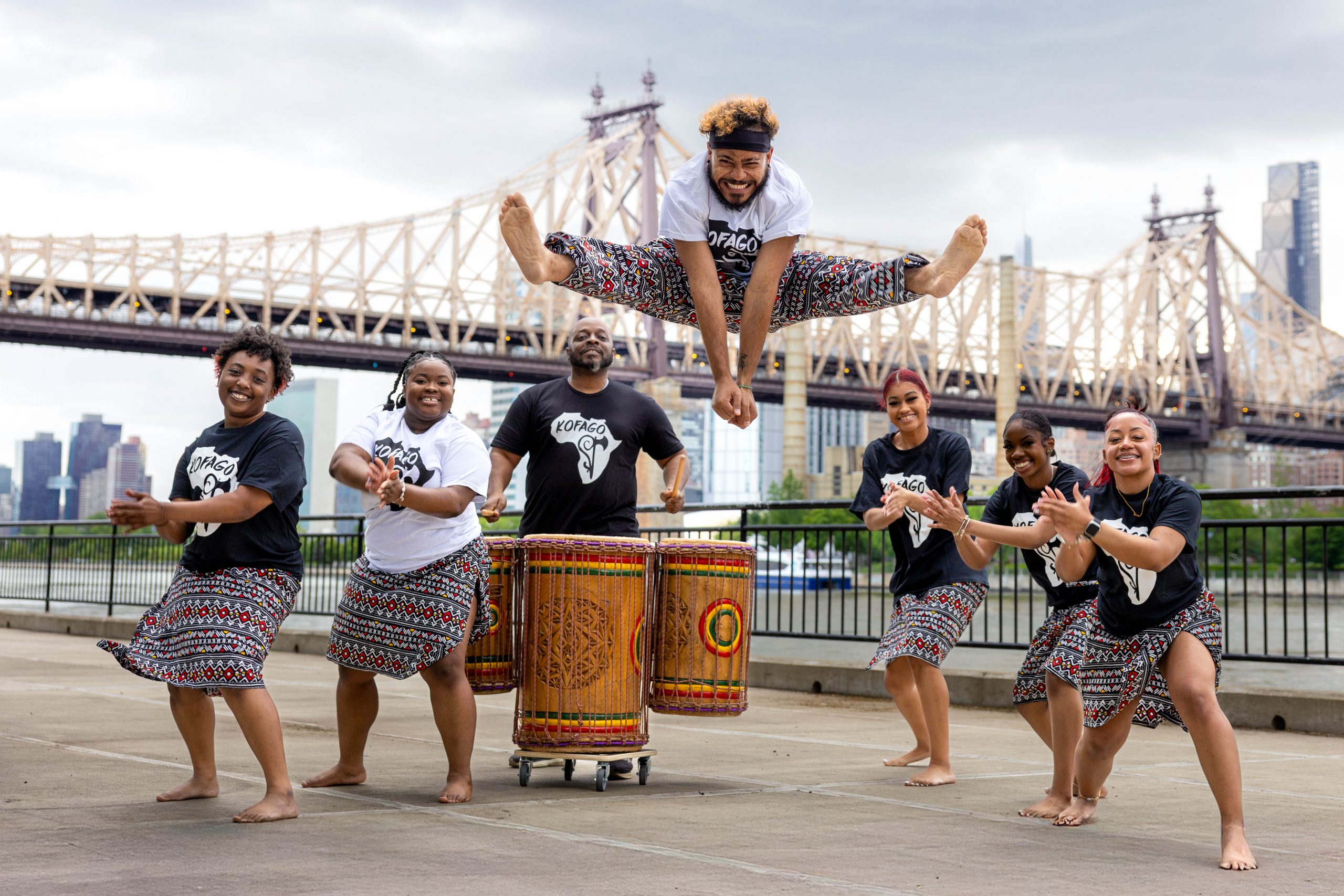 West African dance company in front of Queensborough Bridge. One dancer jump split in air while others clap and play drums behind him.