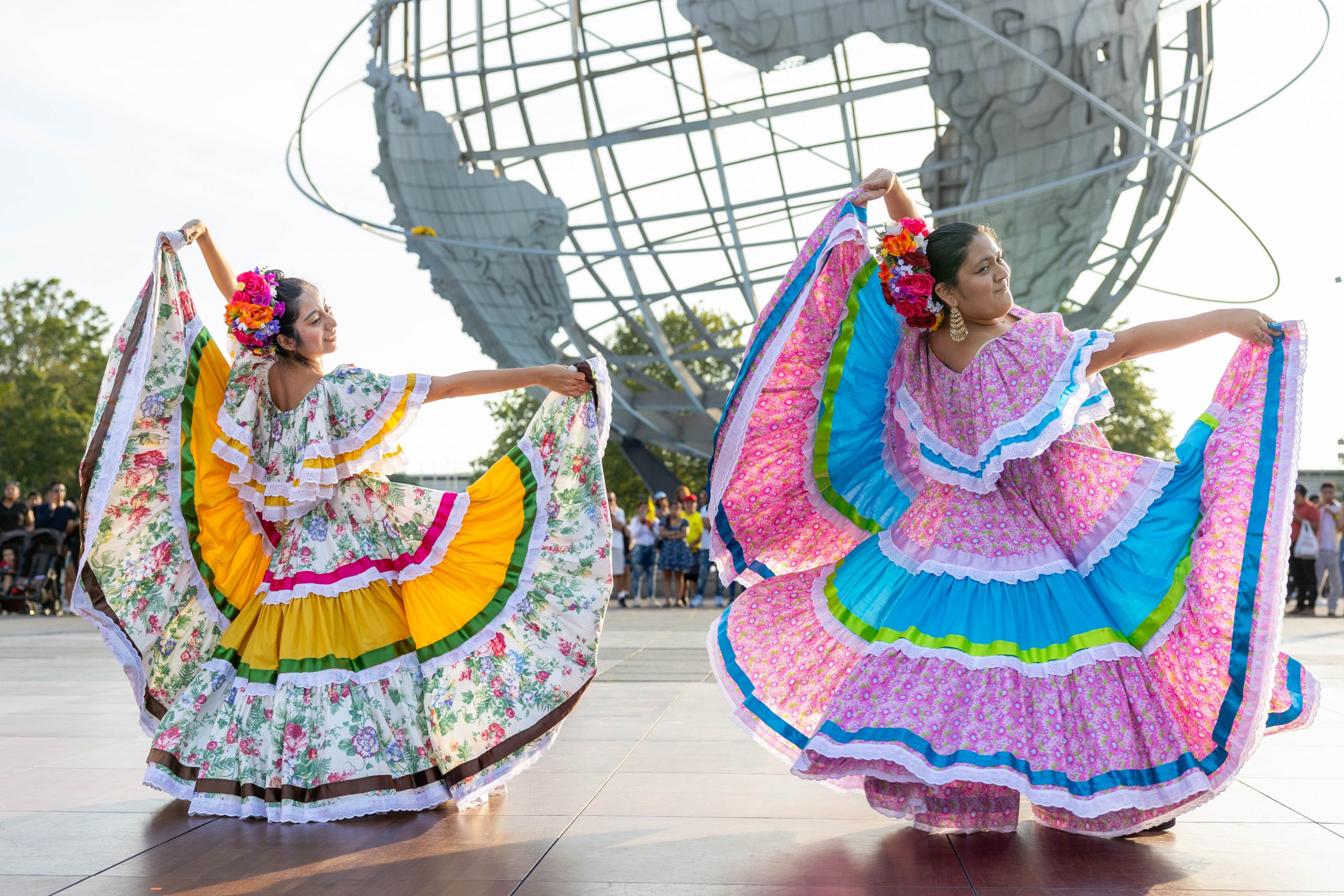 Two Mexican folkloric dancers holding large colorful skirts, Unisphere in background